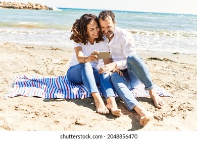 Middle age hispanic couple make selfie by the smartphone sitting on the towel at the beach. - Powered by Shutterstock