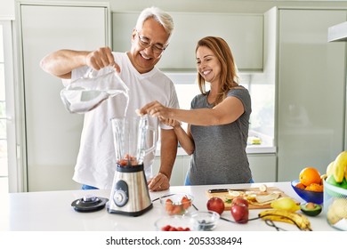 Middle Age Hispanic Couple Pouring Water On Mixer Machine Cooking Smoothie At The Kitchen.