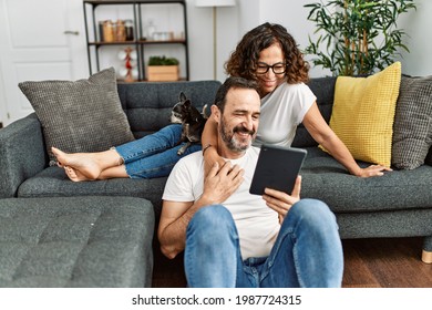 Hispanisches Paar im Mittelalter, das glücklich lächelt und Touchpad benutzt. Sitzen Sie mit Hunden zu Hause auf dem Sofa.