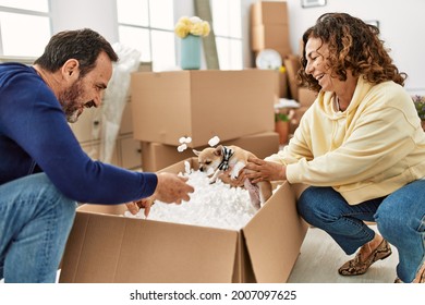 Middle Age Hispanic Couple Smiling Happy Playing With Dog And Foam Package At New Home.