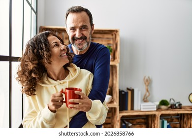 Hispanische Paare mittleren Alters lächeln glücklich und trinken Kaffee. Man lehnt sich zu Hause auf das Fenster.
