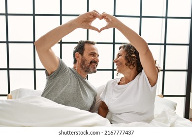 Middle Age Hispanic Couple Smiling Happy Making Heart Symbol With Hands. Sitting On The Bed At Home.