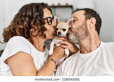 Middle Age Hispanic Couple Smiling Happy Sitting On The Sofa Kissing Dog At Home.