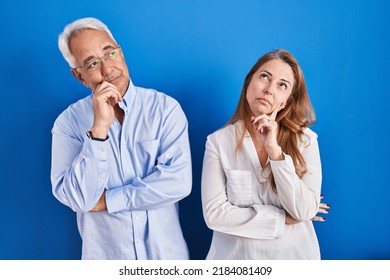 Middle age hispanic couple standing over blue background with hand on chin thinking about question, pensive expression. smiling with thoughtful face. doubt concept. 