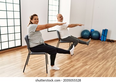 Middle Age Hispanic Couple Stretching Using Chair At Sport Center.