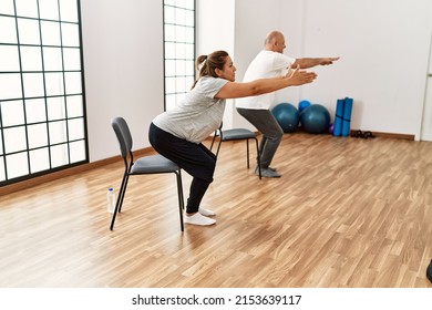 Middle Age Hispanic Couple Stretching Using Chair At Sport Center.