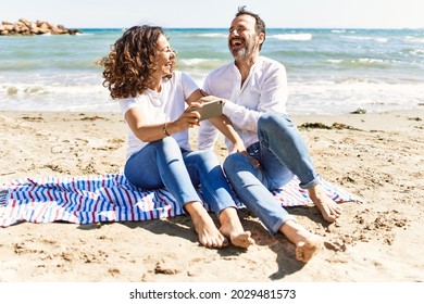 Middle Age Hispanic Couple Using Smartphone Sitting On The Towel At The Beach.