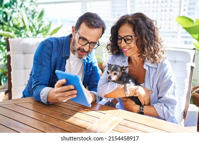 Middle age hispanic couple using touchpad sitting on table with dog at terrace