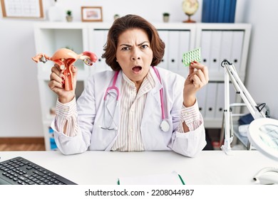 Middle Age Hispanic Doctor Woman Holding Anatomical Female Genital Organ And Birth Control Pills In Shock Face, Looking Skeptical And Sarcastic, Surprised With Open Mouth 