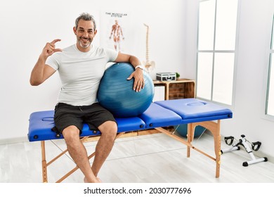 Middle age hispanic man at pain recovery clinic holding pilates ball smiling and confident gesturing with hand doing small size sign with fingers looking and the camera. measure concept. 