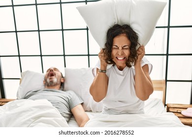 Middle Age Hispanic Man Snoring And Woman Angry Covering Ears With Pillow At Bedroom.