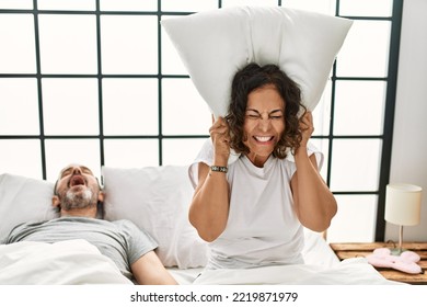 Middle Age Hispanic Man Snoring And Woman Angry Covering Ears With Pillow At Bedroom.