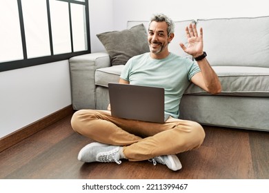 Middle Age Hispanic Man Using Laptop Sitting On The Floor At The Living Room Waiving Saying Hello Happy And Smiling, Friendly Welcome Gesture 