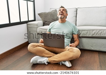 Middle age hispanic man using laptop sitting on the floor at the living room angry and mad screaming frustrated and furious, shouting with anger. rage and aggressive concept. 