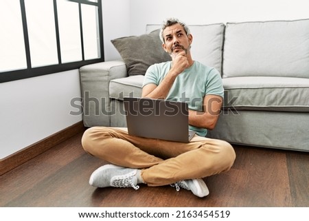 Middle age hispanic man using laptop sitting on the floor at the living room with hand on chin thinking about question, pensive expression. smiling with thoughtful face. doubt concept. 