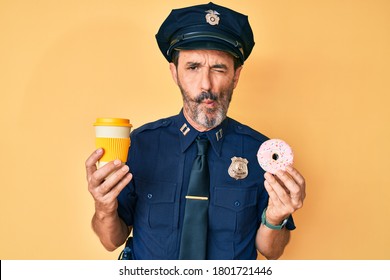 Middle age hispanic man wearing police uniform eating take away coffee and donut making fish face with mouth and squinting eyes, crazy and comical. 
