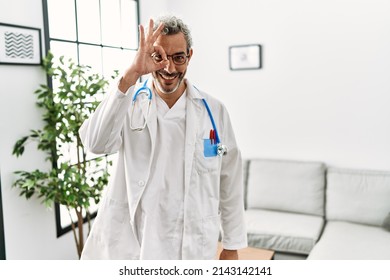 Middle Age Hispanic Man Wearing Doctor Uniform And Stethoscope At Waiting Room Doing Ok Gesture With Hand Smiling, Eye Looking Through Fingers With Happy Face. 