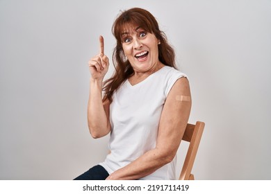 Middle Age Hispanic Woman Getting Vaccine Showing Arm With Band Aid Pointing Finger Up With Successful Idea. Exited And Happy. Number One. 