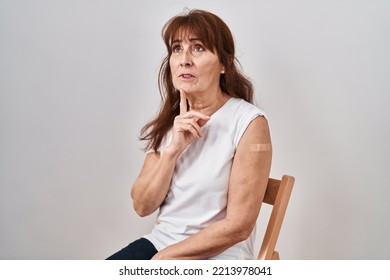 Middle Age Hispanic Woman Getting Vaccine Showing Arm With Band Aid Thinking Concentrated About Doubt With Finger On Chin And Looking Up Wondering 