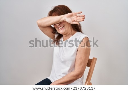 Middle age hispanic woman getting vaccine showing arm with band aid smiling cheerful playing peek a boo with hands showing face. surprised and exited 