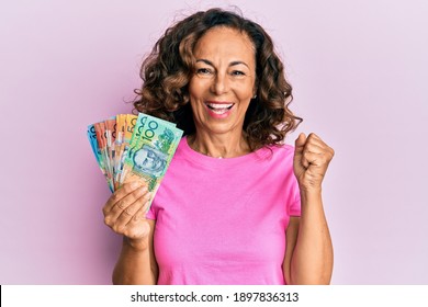 Middle age hispanic woman holding australian dollars screaming proud, celebrating victory and success very excited with raised arm 