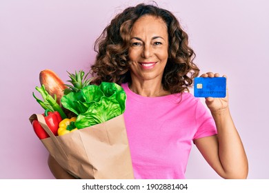 Middle age hispanic woman holding groceries and credit card smiling with a happy and cool smile on face. showing teeth.  - Powered by Shutterstock