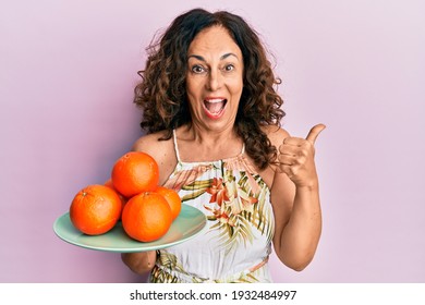 Middle Age Hispanic Woman Holding Plate With Fresh Oranges Pointing Thumb Up To The Side Smiling Happy With Open Mouth 