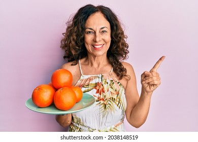 Middle Age Hispanic Woman Holding Plate With Fresh Oranges Smiling Happy Pointing With Hand And Finger To The Side 