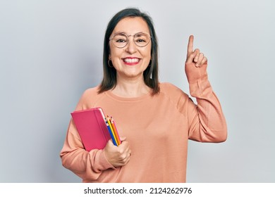 Middle Age Hispanic Woman Holding Book And Color Pencils Surprised With An Idea Or Question Pointing Finger With Happy Face, Number One 