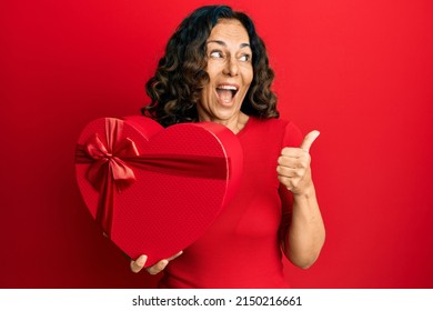 Middle Age Hispanic Woman Holding Valentine Day Gift Pointing Thumb Up To The Side Smiling Happy With Open Mouth 