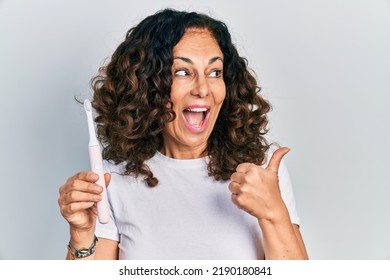 Middle Age Hispanic Woman Holding Electric Toothbrush Pointing Thumb Up To The Side Smiling Happy With Open Mouth 