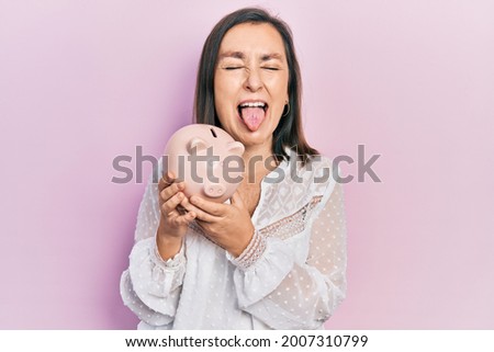 Middle age hispanic woman holding piggy bank sticking tongue out happy with funny expression. 