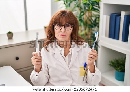 Middle age hispanic woman holding ordinary toothbrush and electric toothbrush relaxed with serious expression on face. simple and natural looking at the camera. 