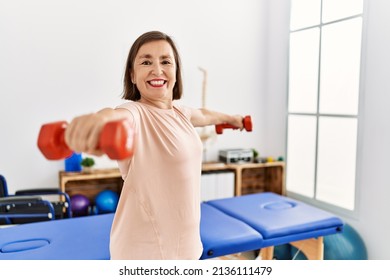 Middle Age Hispanic Woman Lifting Weights At Physiotherapy Clinic