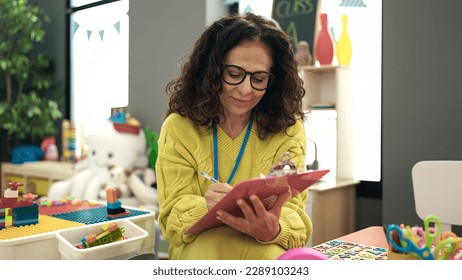 Middle age hispanic woman preschool teacher smiling confident writing on document at kindergarten - Powered by Shutterstock