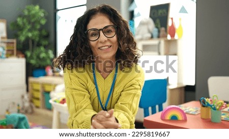 Middle age hispanic woman preschool teacher smiling confident sitting on chair at kindergarten