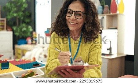 Middle age hispanic woman preschool teacher smiling confident writing on document at kindergarten
