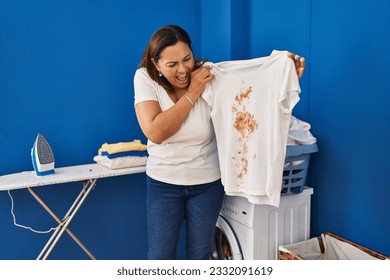 Middle age hispanic woman putting dirty laundry into washing machine angry and mad screaming frustrated and furious, shouting with anger. rage and aggressive concept. 