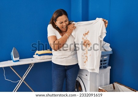 Middle age hispanic woman putting dirty laundry into washing machine angry and mad screaming frustrated and furious, shouting with anger. rage and aggressive concept. 