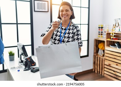 Middle Age Hispanic Woman Shopkeeper Holding Shopping Bag At Clothing Store