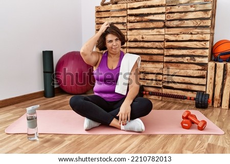 Middle age hispanic woman sitting on training mat at the gym confuse and wondering about question. uncertain with doubt, thinking with hand on head. pensive concept. 