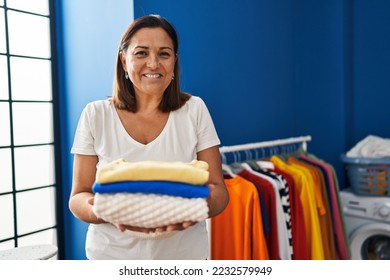 Middle age hispanic woman smiling confident holding folded clothes at laundry room - Powered by Shutterstock