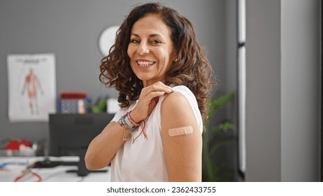 Middle age hispanic woman smiling confident standing with band aid on arm at clinic
