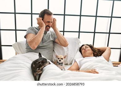 Middle Age Hispanic Woman Snoring And Man Angry Covering Ears At Bedroom.