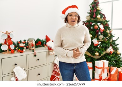 Middle Age Hispanic Woman Standing By Christmas Tree With Hand On Stomach Because Indigestion, Painful Illness Feeling Unwell. Ache Concept. 