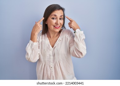 Middle Age Hispanic Woman Standing Over Blue Background Smiling Pointing To Head With Both Hands Finger, Great Idea Or Thought, Good Memory 