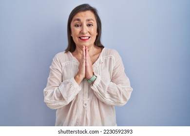 Middle age hispanic woman standing over blue background praying with hands together asking for forgiveness smiling confident.  - Shutterstock ID 2230592393