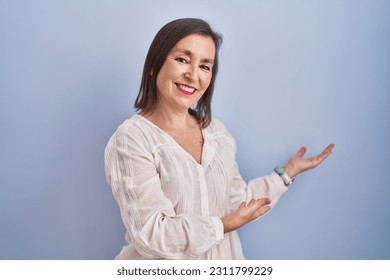 Middle age hispanic woman standing over blue background inviting to enter smiling natural with open hand  - Shutterstock ID 2311799229