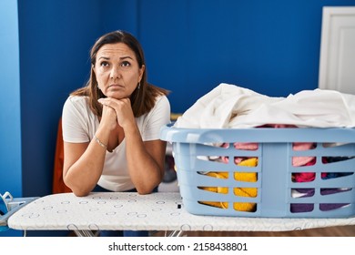 Middle Age Hispanic Woman Tired Leaning On Ironing Board At Laundry Room