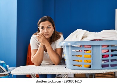 Middle Age Hispanic Woman Tired Leaning On Ironing Board At Laundry Room